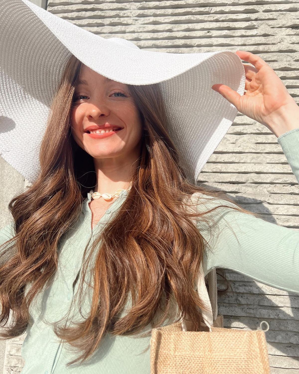 Woman in a wide-brimmed white hat with long, loose brown waves, standing on a balcony in front of a city skyline.