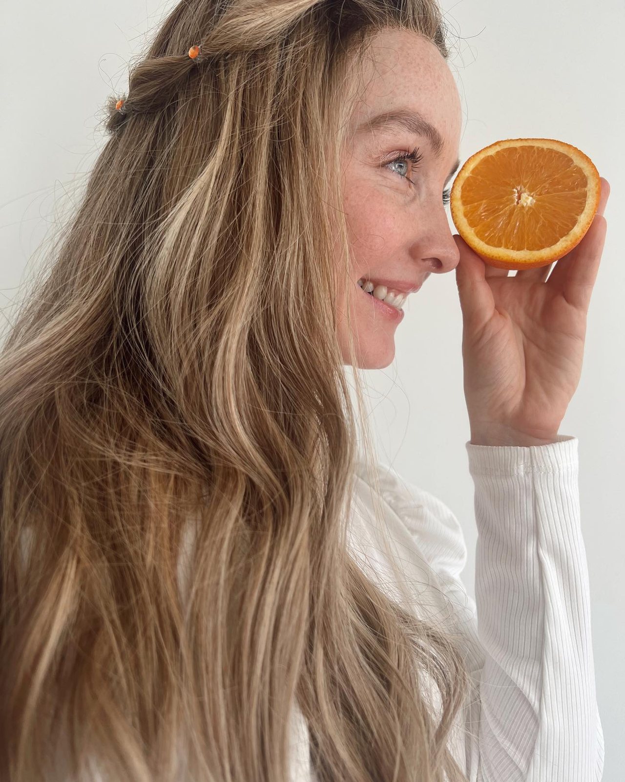 Long, layered blonde waves with face-framing pieces, and a slice of orange held near the chin.