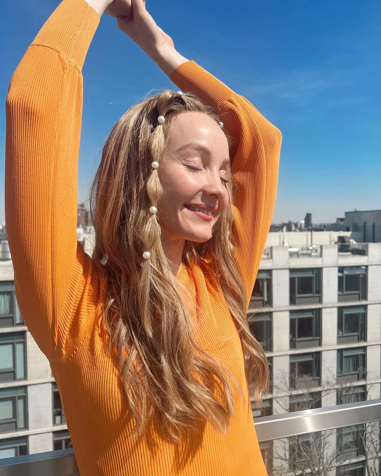 Pearl-studded braids gathered low, with a bright orange sweater and the Manhattan skyline behind.