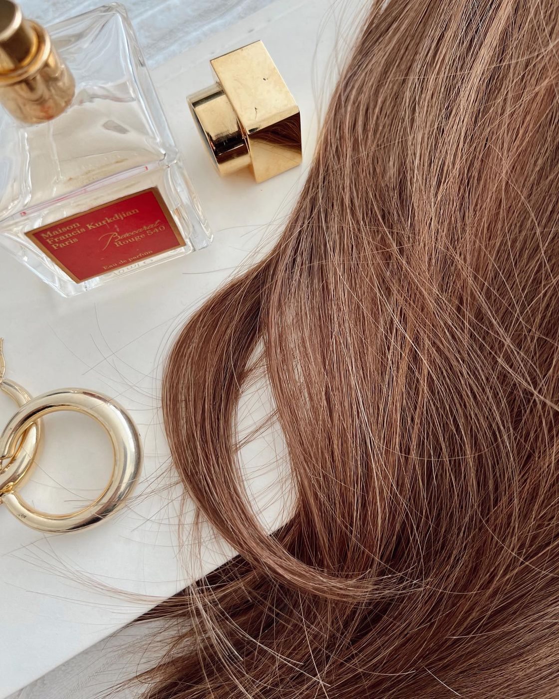 Close-up of auburn waves beside a square glass perfume bottle and gold jewelry.